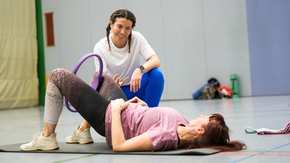 Zwei Frauen in Sportkleidung trainieren auf einer Matte, eine unterstützt die andere mit einem Fitnessgerät.