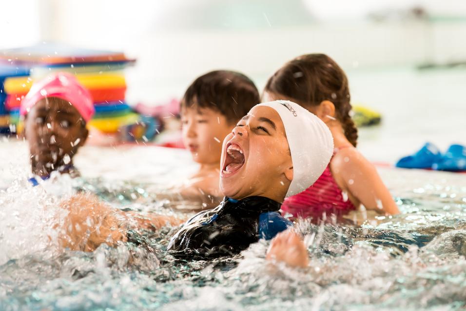Vier Kinder spielen im Wasser eines Schwimmbads, ein Junge lacht laut und spritzt Wasser mit seinen Händen.