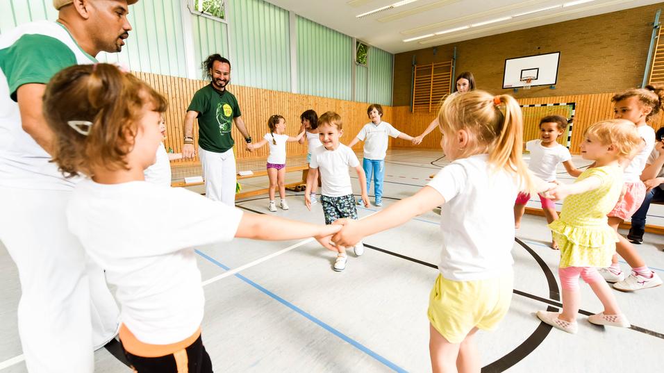 Gruppe von Kindern in T-Shirts und Erwachsenen in einer Turnhalle, die gemeinsam einen Kreis bilden und spielen.