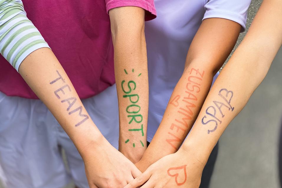 Vier Arme liegen übereinander, mit farbiger Schrift: „TEAM“, „SPORT“, „ZUSAMMEN“, „SPASS“ auf der Haut.