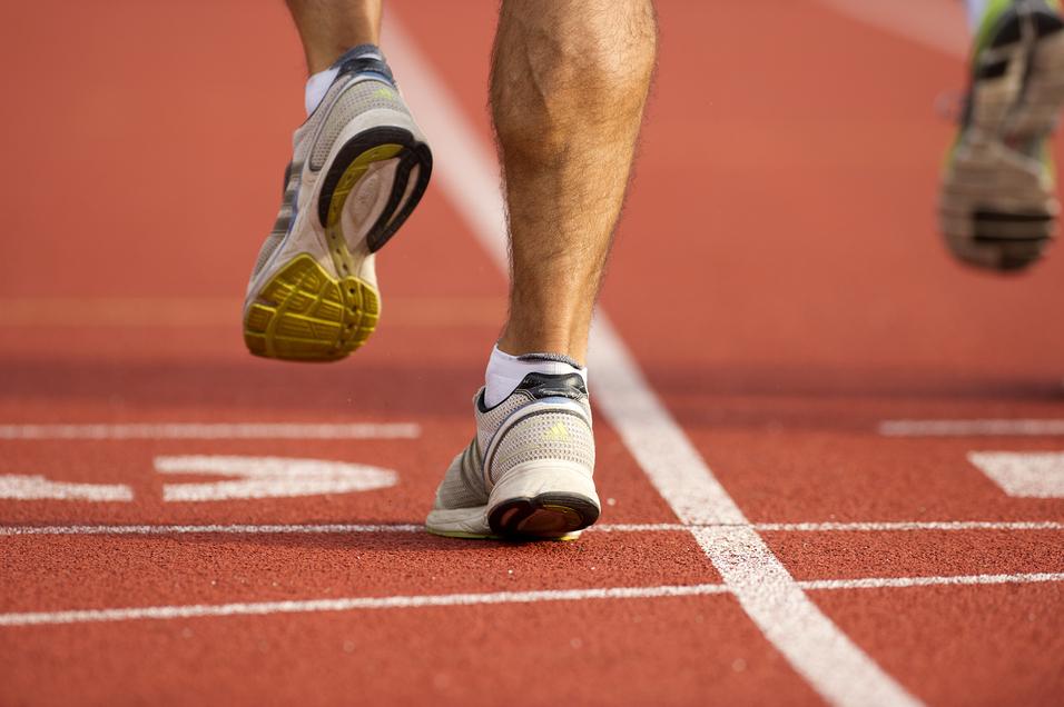 Nahaufnahme eines Läufers auf einem roten Laufbahnbelag, der gerade aufsetzt. Sportschuh mit sichtbarem Profil.