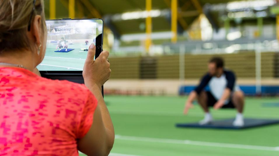 Eine Person filmt einen Mann, der auf einer Matte in der Squat-Position trainiert, in einem Sportzentrum.