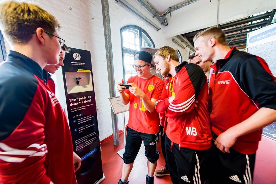 Gruppe junger Sportler in roten Trikots schaut auf ein Smartphone und diskutiert an einem Informationsstand.