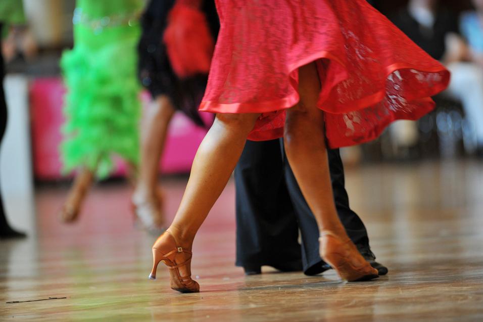 Tanzende Beine in eleganten Schuhen, während sich ein rotes Kleid dreht, vor unscharfem Hintergrund.
