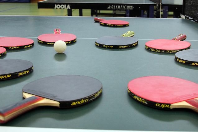 Mehrere Tischtennisschläger in verschiedenen Farben und ein Tischtennisball liegen auf einer grünen Tischtennisplatte.