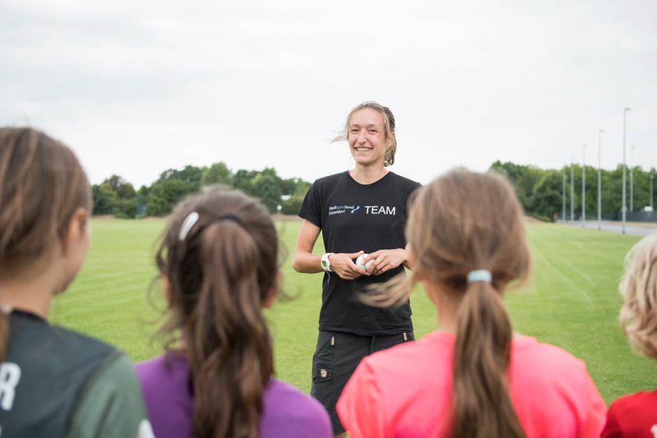 Trainerin spricht mit jungen Sportlerinnen auf einem grünen Fußballfeld unter bewölkten Himmel.