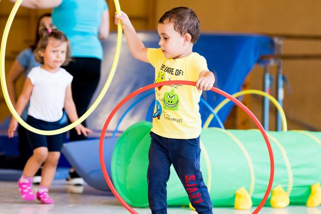 Zwei Kinder spielen in einer Turnhalle mit hula-hoops und einem grünen Tunnel. Ein Kind hält zwei Reifen in der Hand.