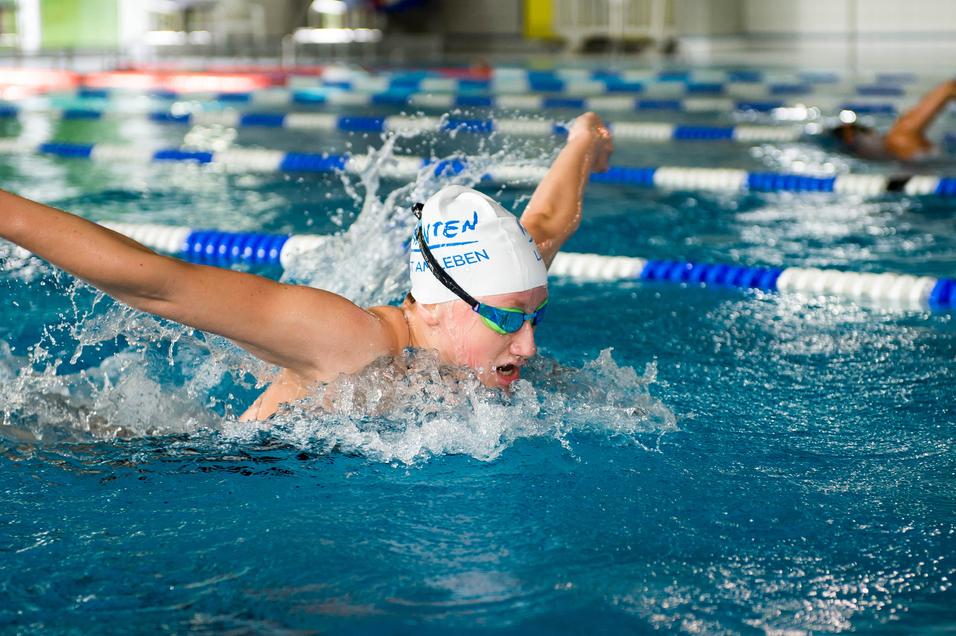 Junge Schwimmerin im Wettkampf, die im Wasser den Schmetterlingsstil ausführt, mit spritzenden Wassertropfen.