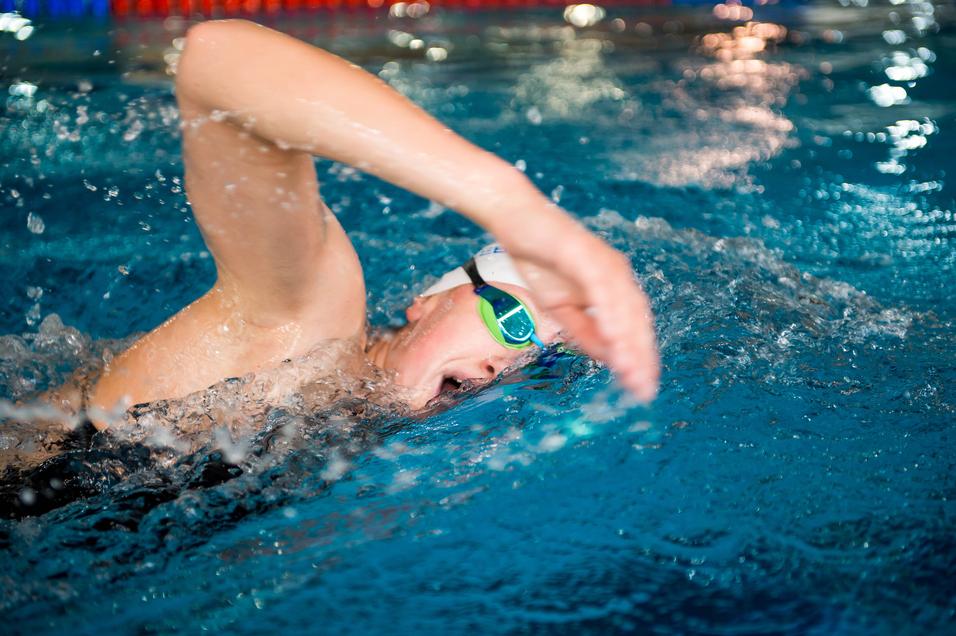 Junge schwimmt mit Schwimmbrille im Wasser, spritzt Wasser mit einer kraftvollen Armbewegung nach vorne.