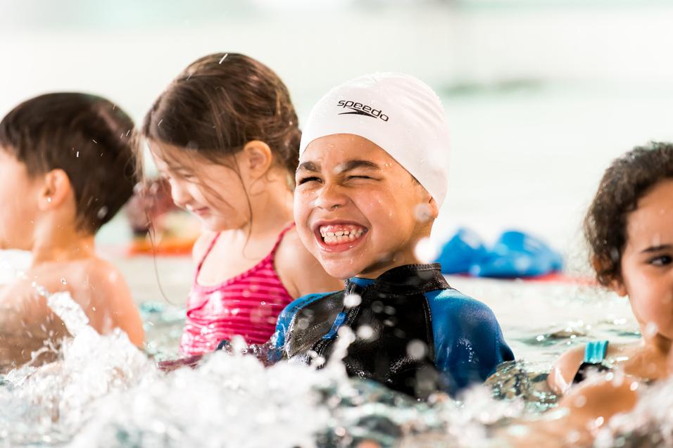 Junge im Schwimmbecken mit weißer Badekappe lächelt, umgeben von weiteren Kindern und spritzendem Wasser.