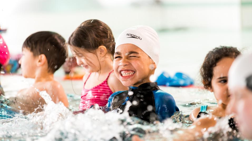 Junge im Schwimmbecken mit weißer Badekappe lächelt, umgeben von weiteren Kindern und spritzendem Wasser.